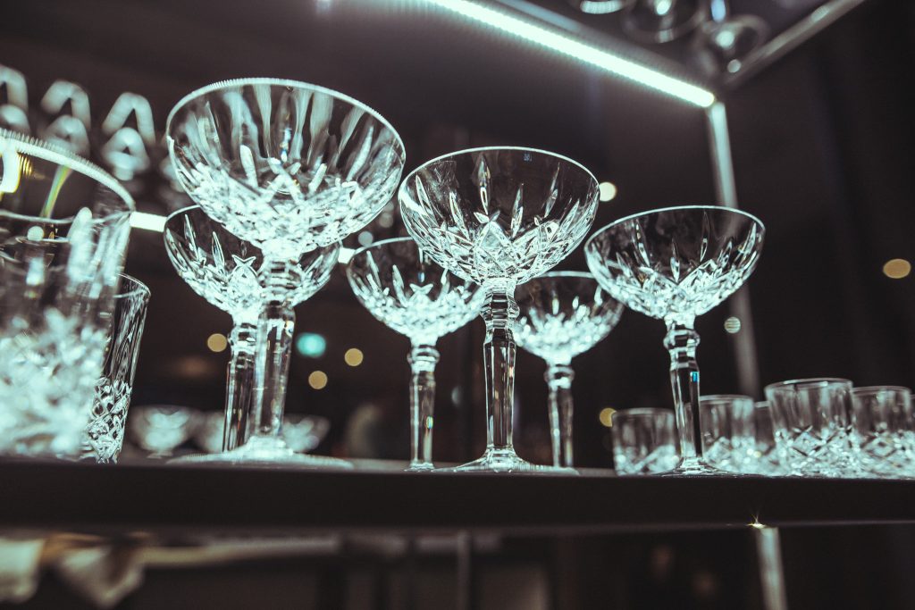 © Sergiu Andrés Crystal stemware arranged on a bar shelf with warm lighting and reflections on the glass surfaces.