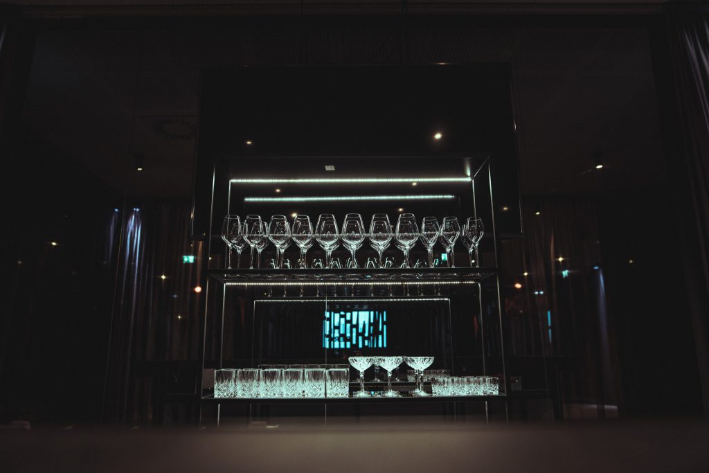 © Sergiu Andrés Rows of stemmed wine glasses hang upside down on a backlit glass shelf above a bar in a dim venue.