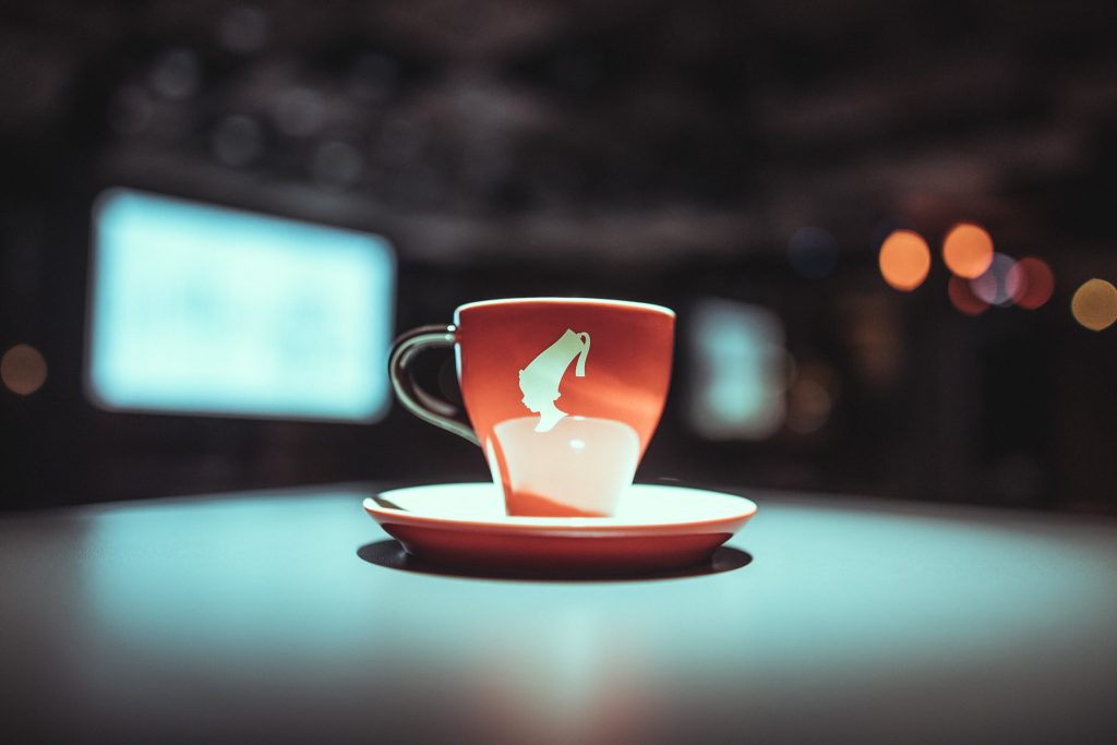 © Sergiu Andrés Red ceramic cup with white interior and a white design on the side sits on a matching saucer on a teal-lit surface, with blurred city lights in the background, creating bokeh.