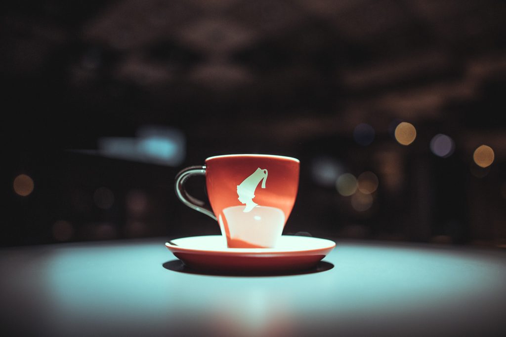 © Sergiu Andrés Red teacup with white design on a matching saucer, set on a teal table with blurred city lights in the background.