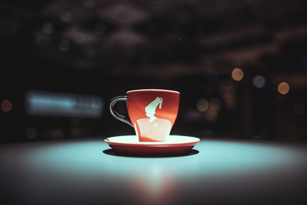 © Sergiu Andrés Red ceramic cup with a white graphic on a matching saucer, placed on a teal table with blurred city lights in the background.