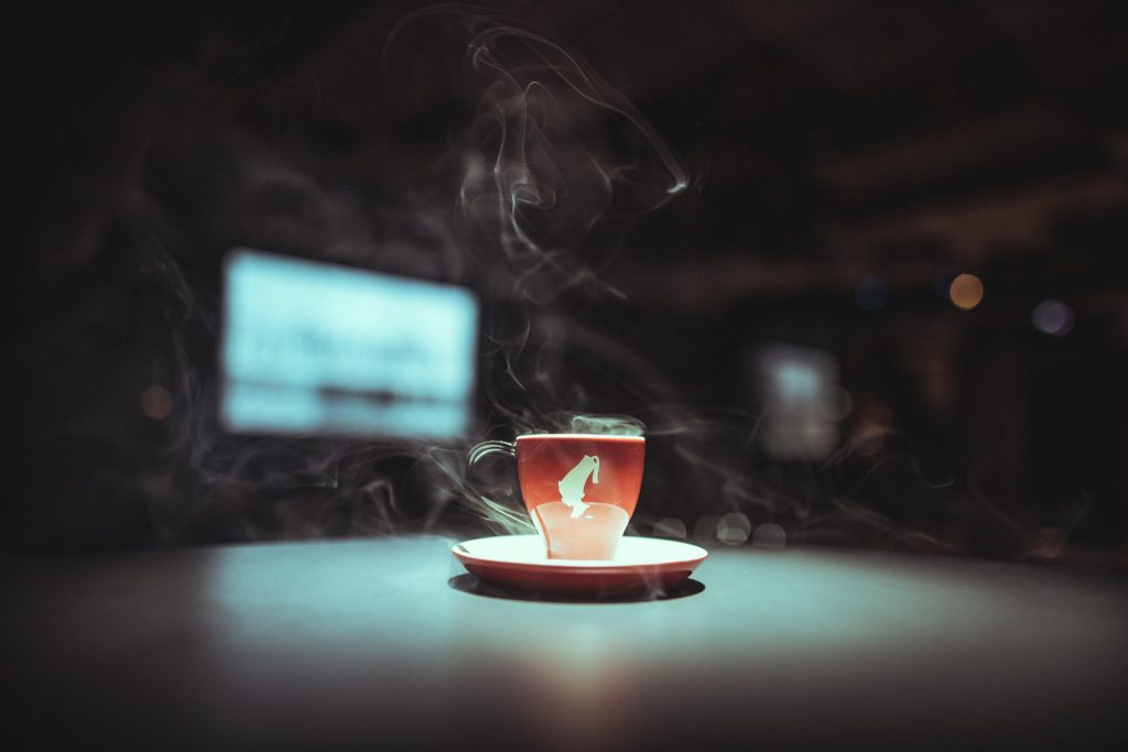 © Sergiu Andrés Red espresso cup on a matching saucer with curling steam against a dark, blurred background, suggesting a hot drink.