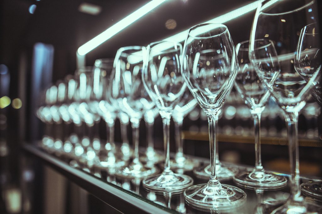 © Sergiu Andrés Row of empty wine glasses lined up on a bar shelf with blurred lights in the background.