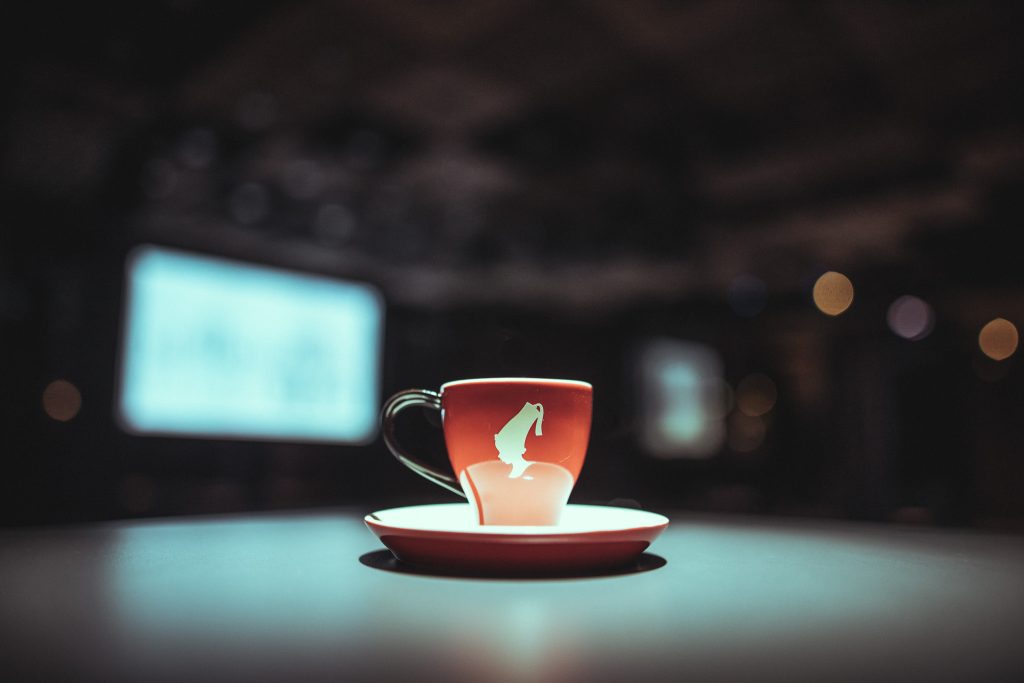 © Sergiu Andrés Red mug with a white horse logo on a matching saucer, set on a table with a blurred dark background and bokeh lights.