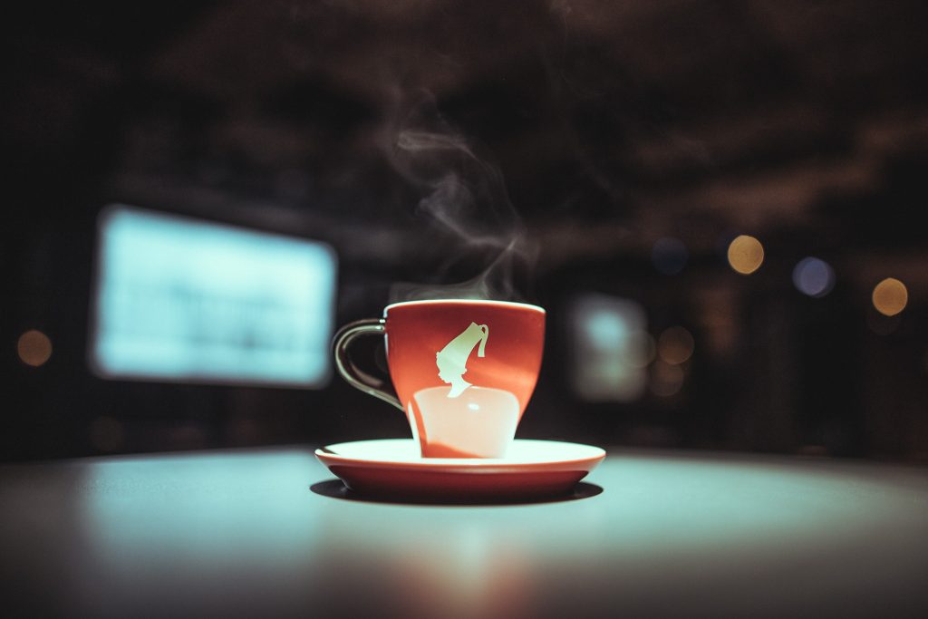 © Sergiu Andrés Red ceramic cup with steam rising on a matching saucer, set on a smooth surface with blurred city lights in the background.