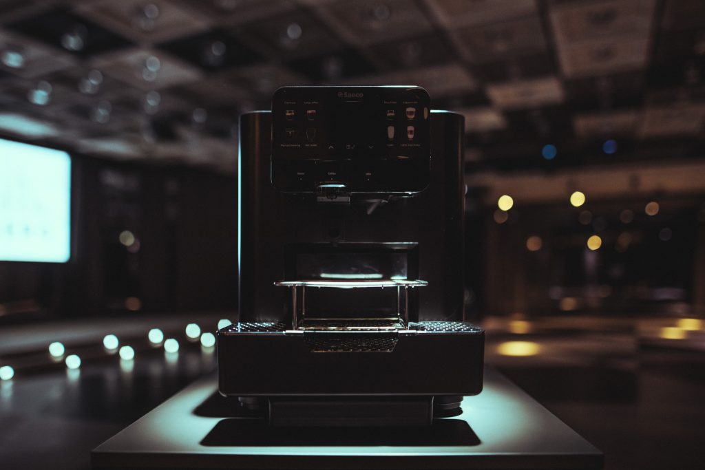 © Sergiu Andrés A dark-lit espresso machine on a counter in a cafe, with a glossy front and control panel visible.