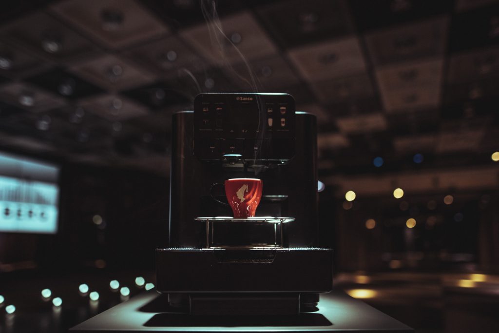 © Sergiu Andrés Close-up of a Saeco espresso machine with a red cup on the drip tray, steam rising in a dim cafe setting.