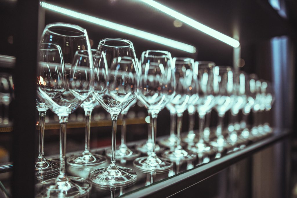 © Sergiu Andrés Row of wine glasses on a bar shelf with bright horizontal lights blurred in the background.