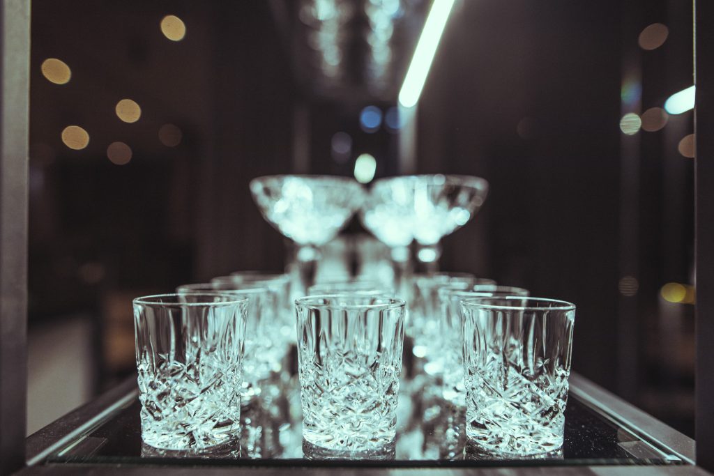 © Sergiu Andrés Row of cut-crystal glasses lined up on a reflective bar surface with soft circular bokeh lights in the background